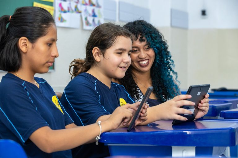 Foto de uma professora e duas alunas mexendo em um celular, simbolizando o estudante bilíngue.